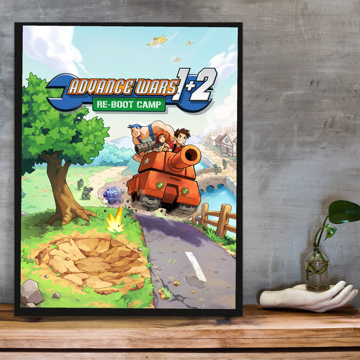 Advance Wars 1+2: Re-Boot Camp Key Art poster in a black frame on a wooden shelf against concrete