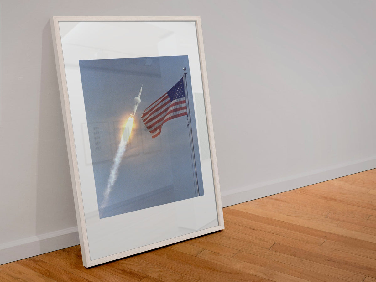 Apollo 11 rocket launch with the US flag poster in a white frame leaning against a wall
