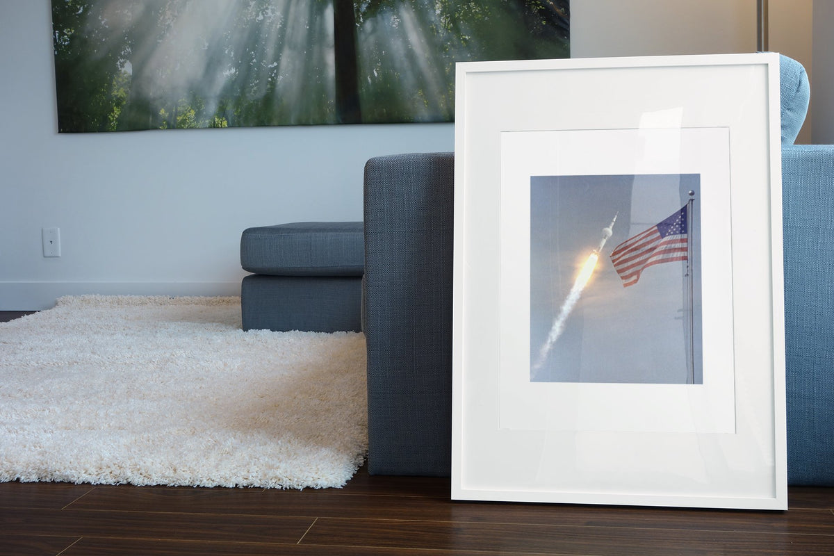 Apollo 11 rocket launch with the US flag poster in a white frame leaning beside a blue sofa