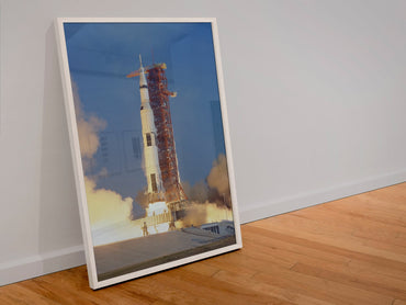 Apollo 11 Saturn V rocket launch poster in a white frame leaning against a wall