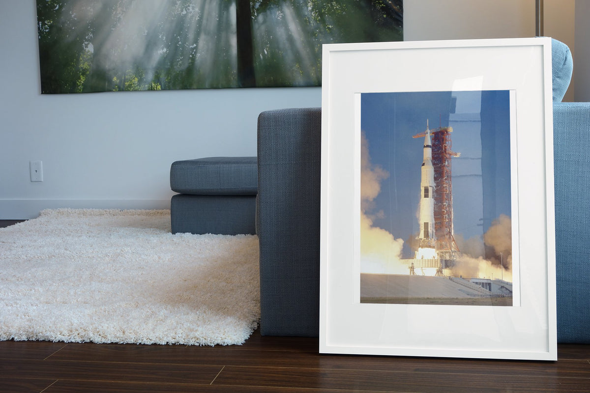 Apollo 11 Saturn V rocket launch poster in a white frame leaning beside a blue sofa