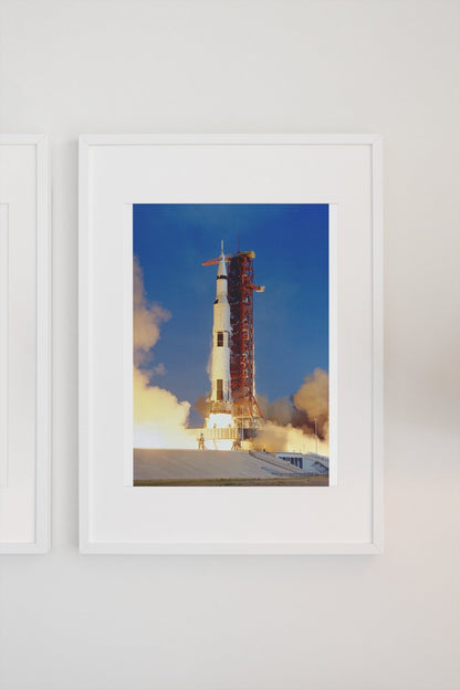 Apollo 11 Saturn V rocket launch poster in a white frame on a light wall