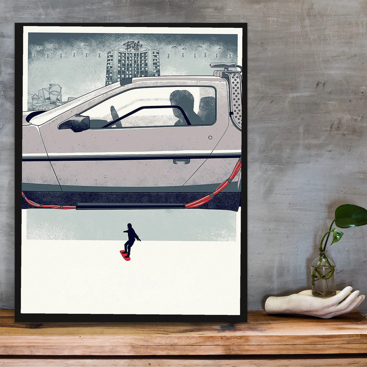 Back to the Future Part II (1989) Movie DeLorean Hover Art poster in a black frame on a wooden shelf against concrete