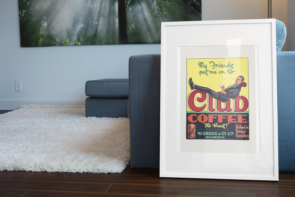 Club Coffee 1920s poster in a white frame leaning beside a blue sofa