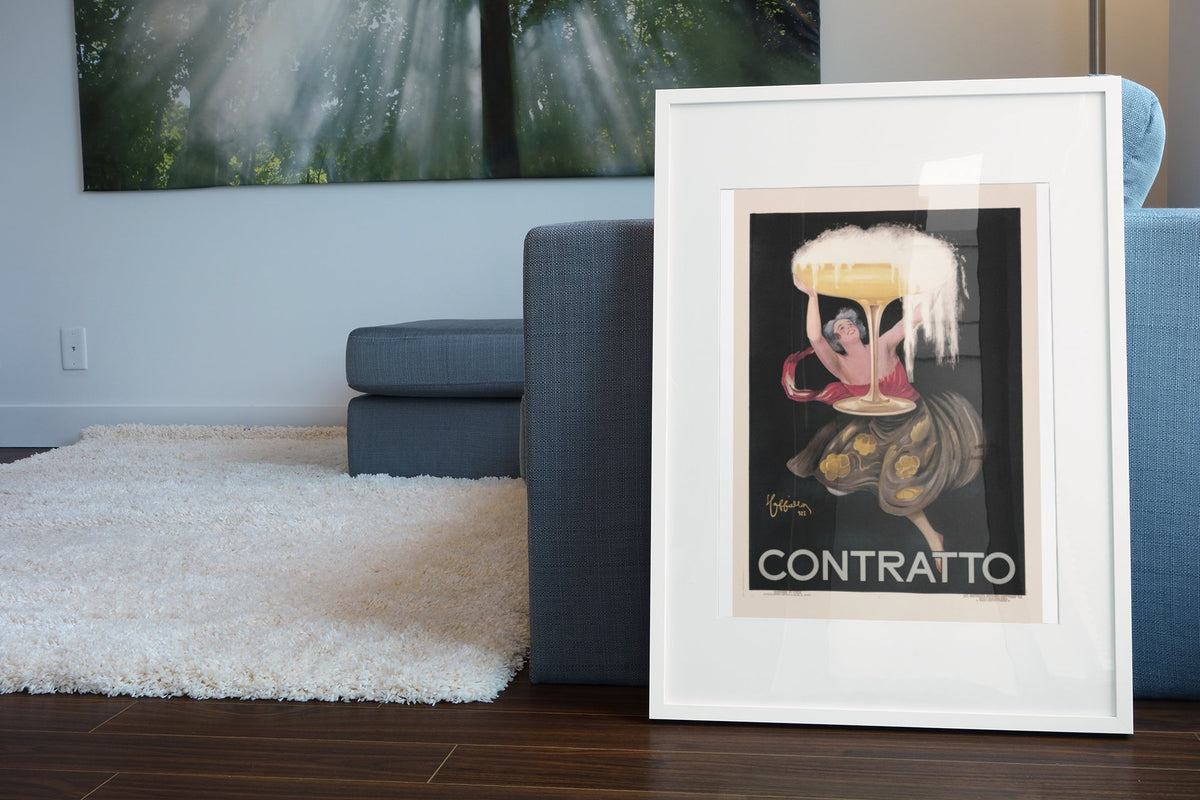 Contratto 1922 champagne by Cappiello poster in a white frame leaning beside a blue sofa