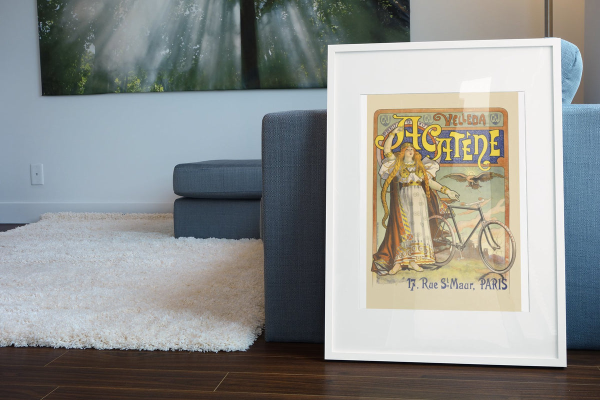 Cycles Acatène Paris bicycle poster in a white frame leaning beside a blue sofa