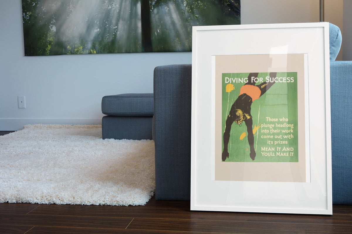 Diving for Success poster in a white frame leaning beside a blue sofa