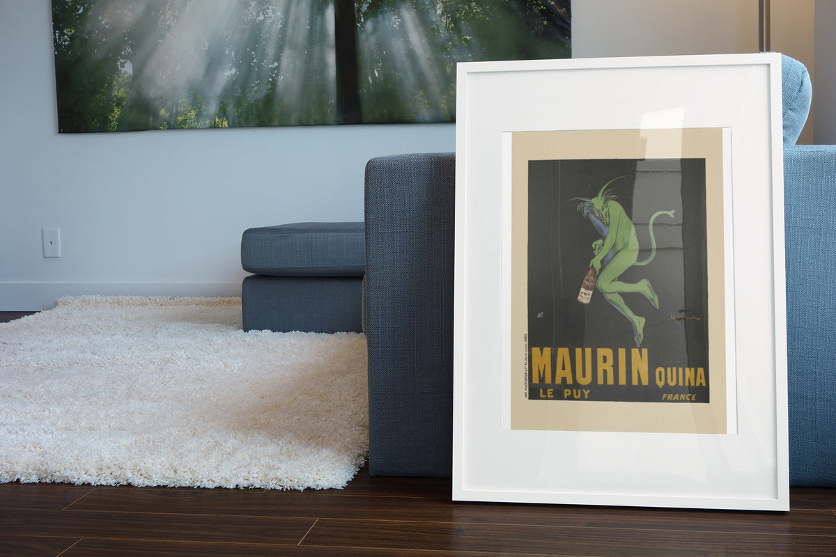 Maurin Quina 1906 green devil by Cappiello poster in a white frame leaning beside a blue sofa