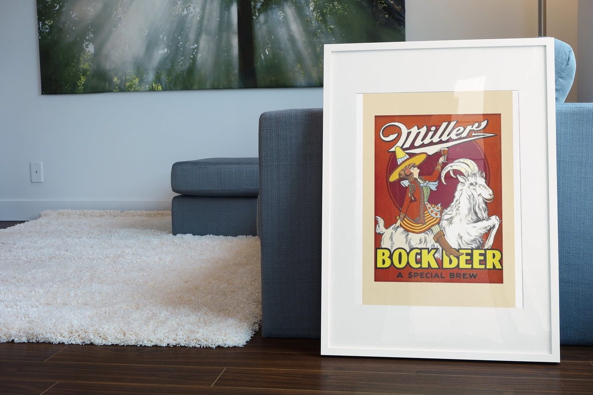 Miller Bock Beer 1930s poster in a white frame leaning beside a blue sofa