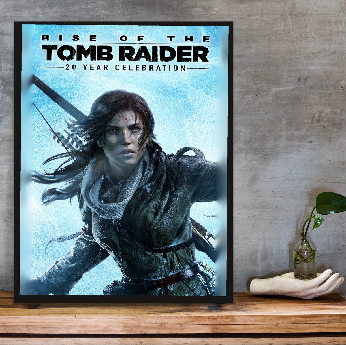 Rise of the Tomb Raider 20 Year Celebration Art poster in a black frame on a wooden shelf against concrete