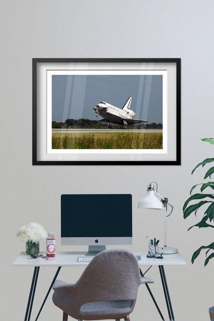 Space Shuttle Atlantis landing poster framed above a desk with a computer