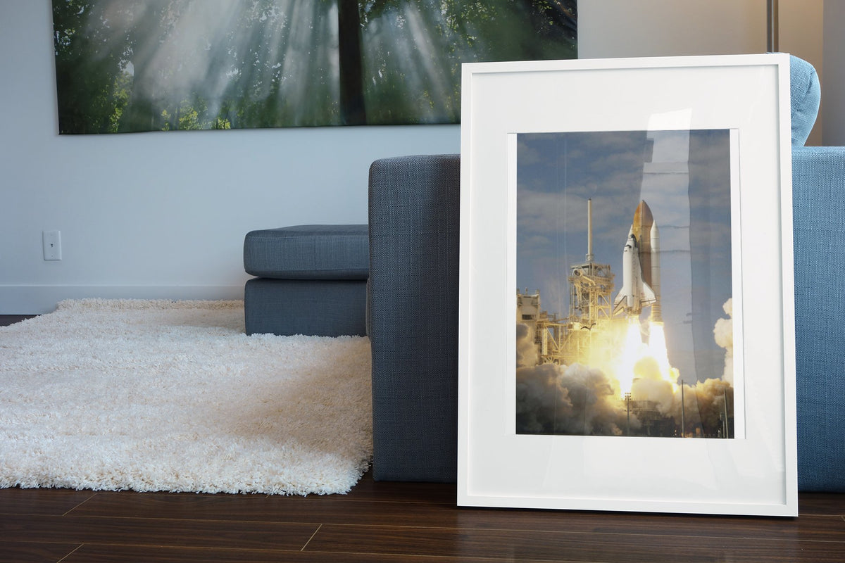 Space Shuttle Atlantis STS-122 launch poster in a white frame leaning beside a blue sofa