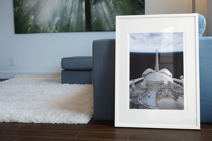 STS-123 Space Shuttle leaving the ISS poster in a white frame leaning beside a blue sofa