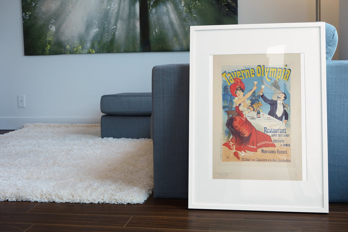 Taverne Olympia 1900 poster in a white frame leaning beside a blue sofa