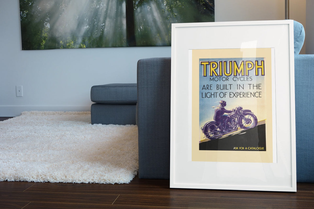 Triumph Motorcycles poster in a white frame leaning beside a blue sofa